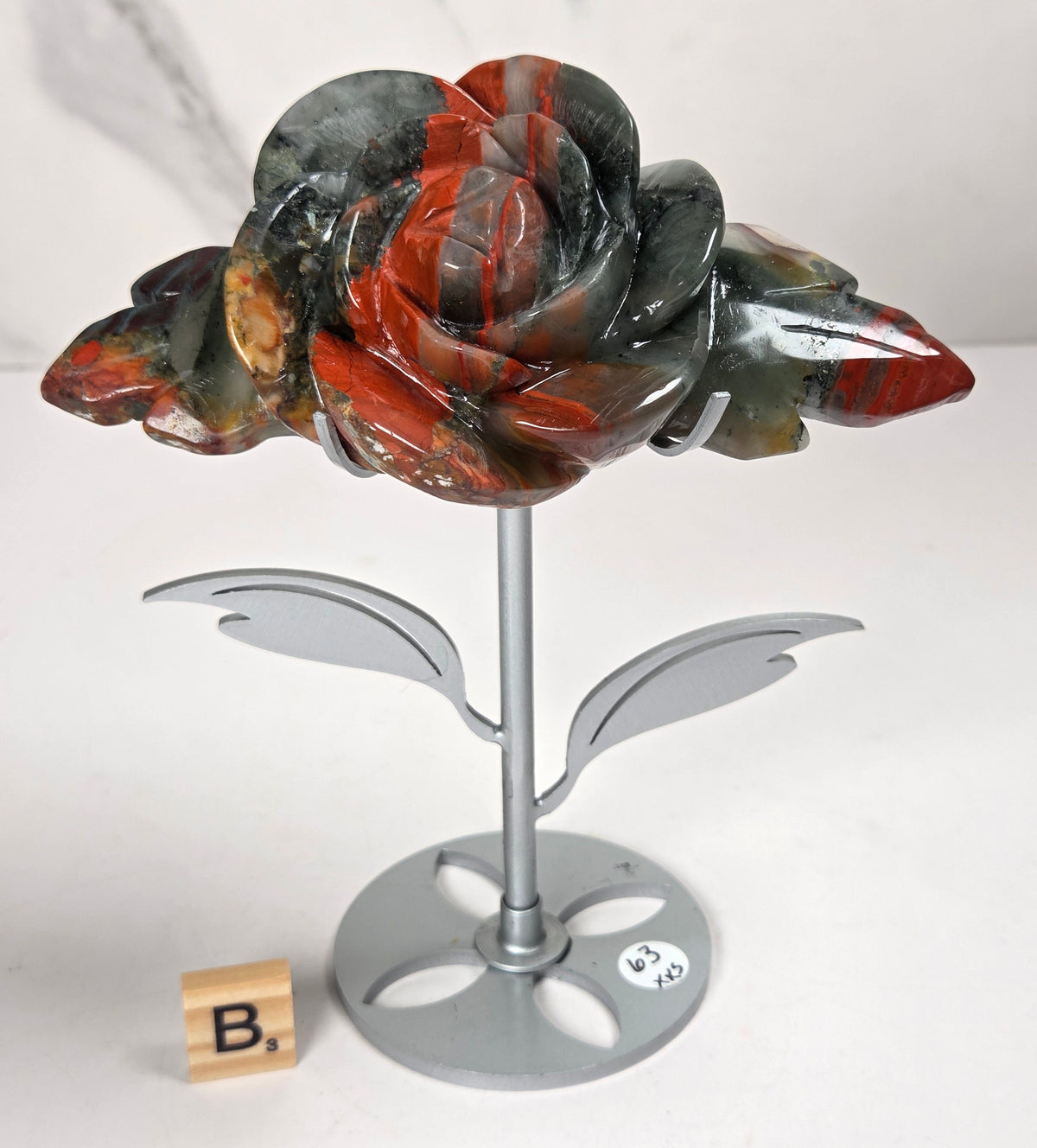 A carved stone rose sculpture with red, brown, and gray marbled petals on a silver metal stem and round base, displayed on a white surface with a small wooden letter tile nearby.