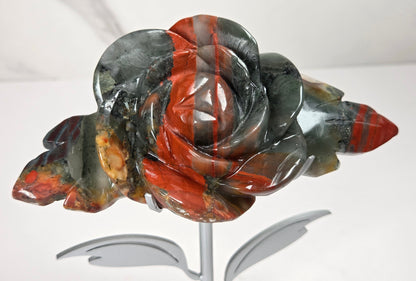 A carved stone rose sculpture with red, brown, and gray marbled petals on a silver metal stem and round base, displayed on a white surface with a small wooden letter tile nearby.