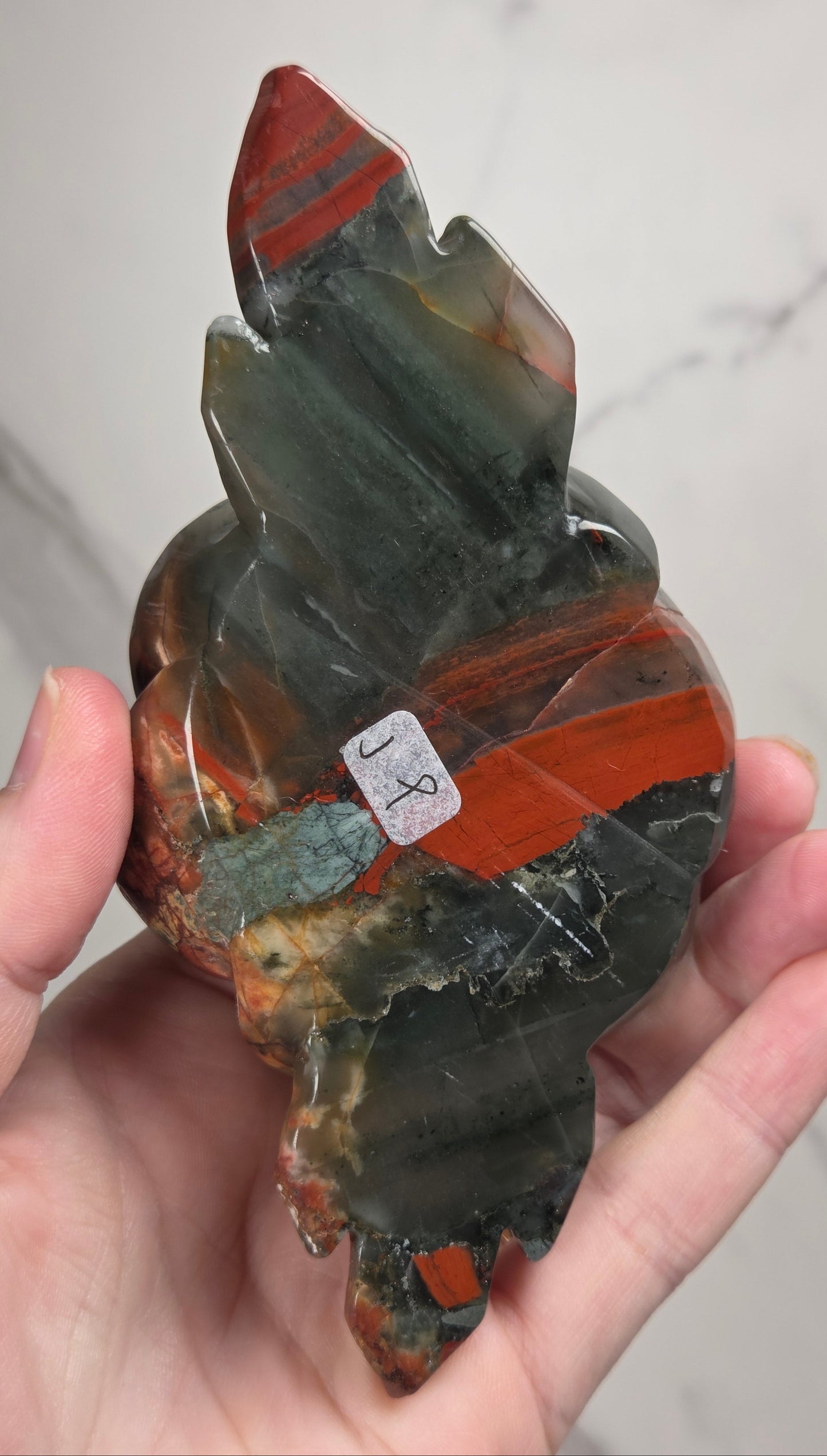 A carved stone rose sculpture with red, brown, and gray marbled petals on a silver metal stem and round base, displayed on a white surface with a small wooden letter tile nearby.