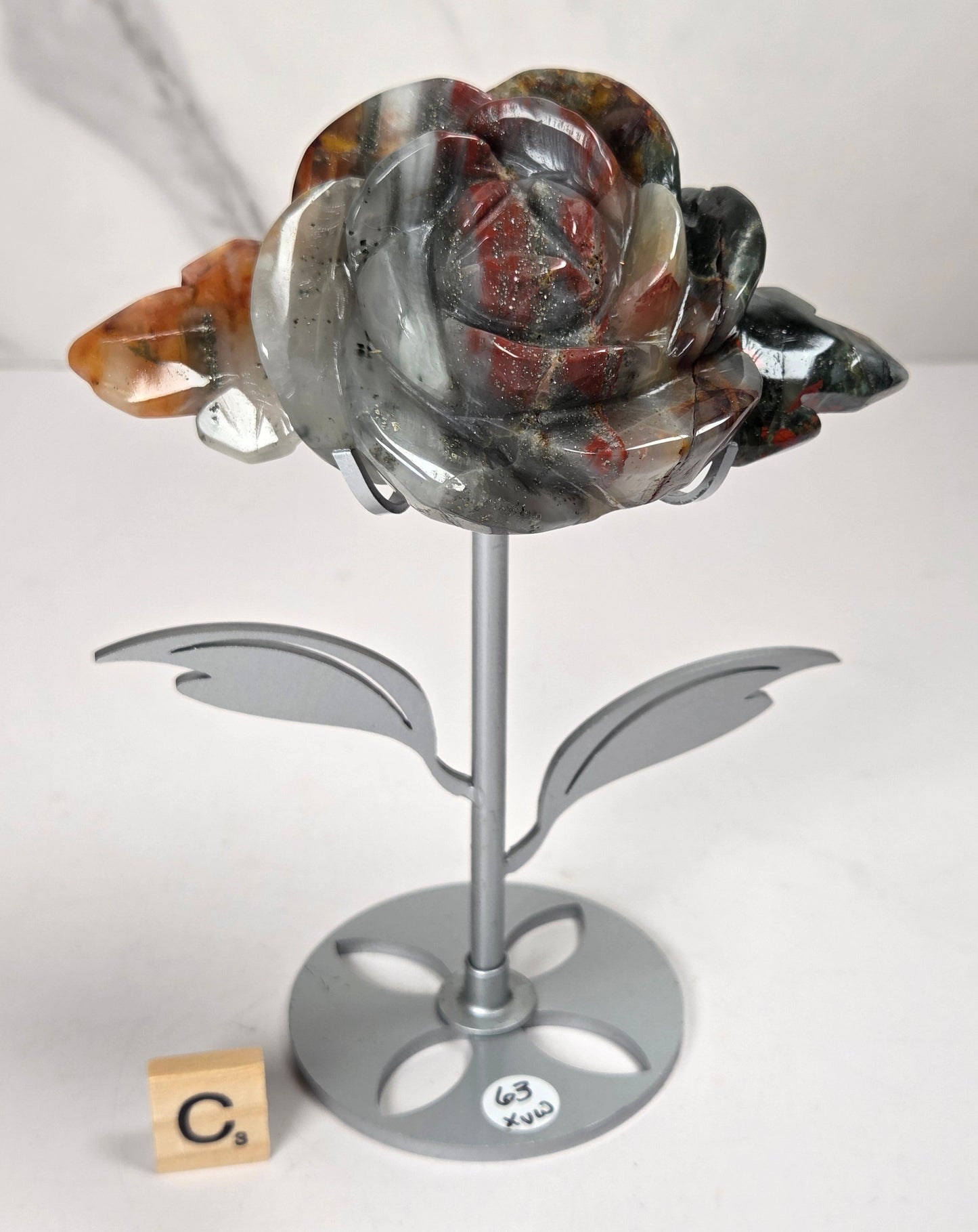 A polished agate rose figurine on a silver metal stand, displayed on a white surface with a marble-patterned background. The agate features natural marbled patterns of gray, black, red, and orange, with a small wooden block displaying 'C.