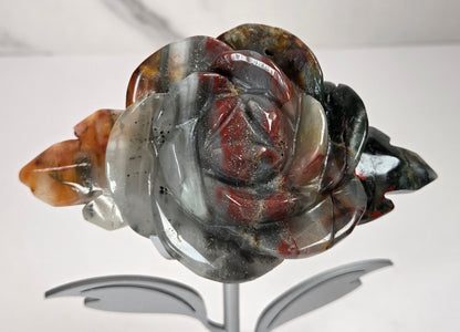 A carved rose-shaped decorative object made from polished agate or jasper stone, displayed on a small metallic stand against a light gray background.