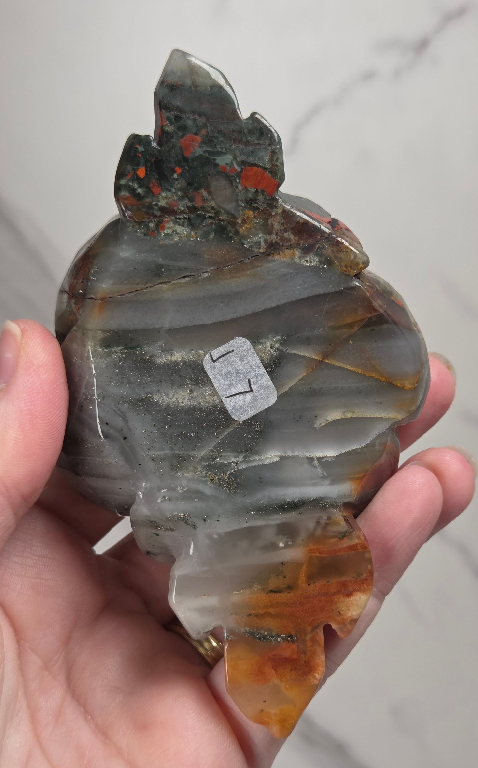 A hand holding a polished agate stone carving with layered bands of gray, white, and amber-brown, featuring small orange-red inclusions near the top.