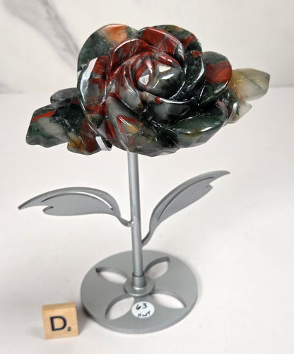 A carved stone rose sculpture with red, brown, and gray marbled petals on a silver metal stem and round base, displayed on a white surface with a small wooden letter tile nearby.