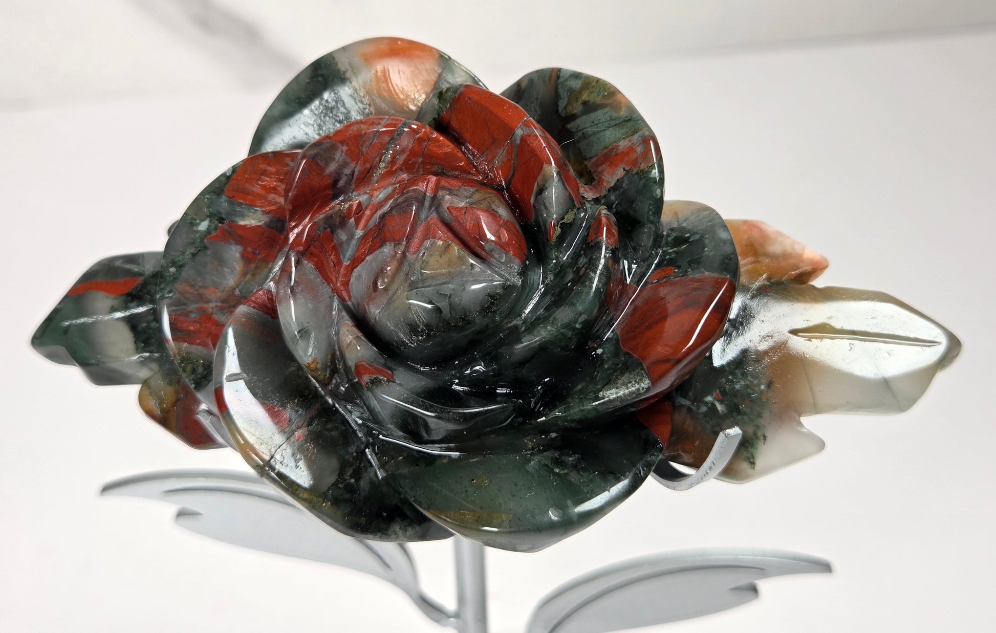 A carved rose-shaped decorative object made from polished stone with swirling bands of dark green, black, and reddish brown, displayed on a silver stand against a plain light background.