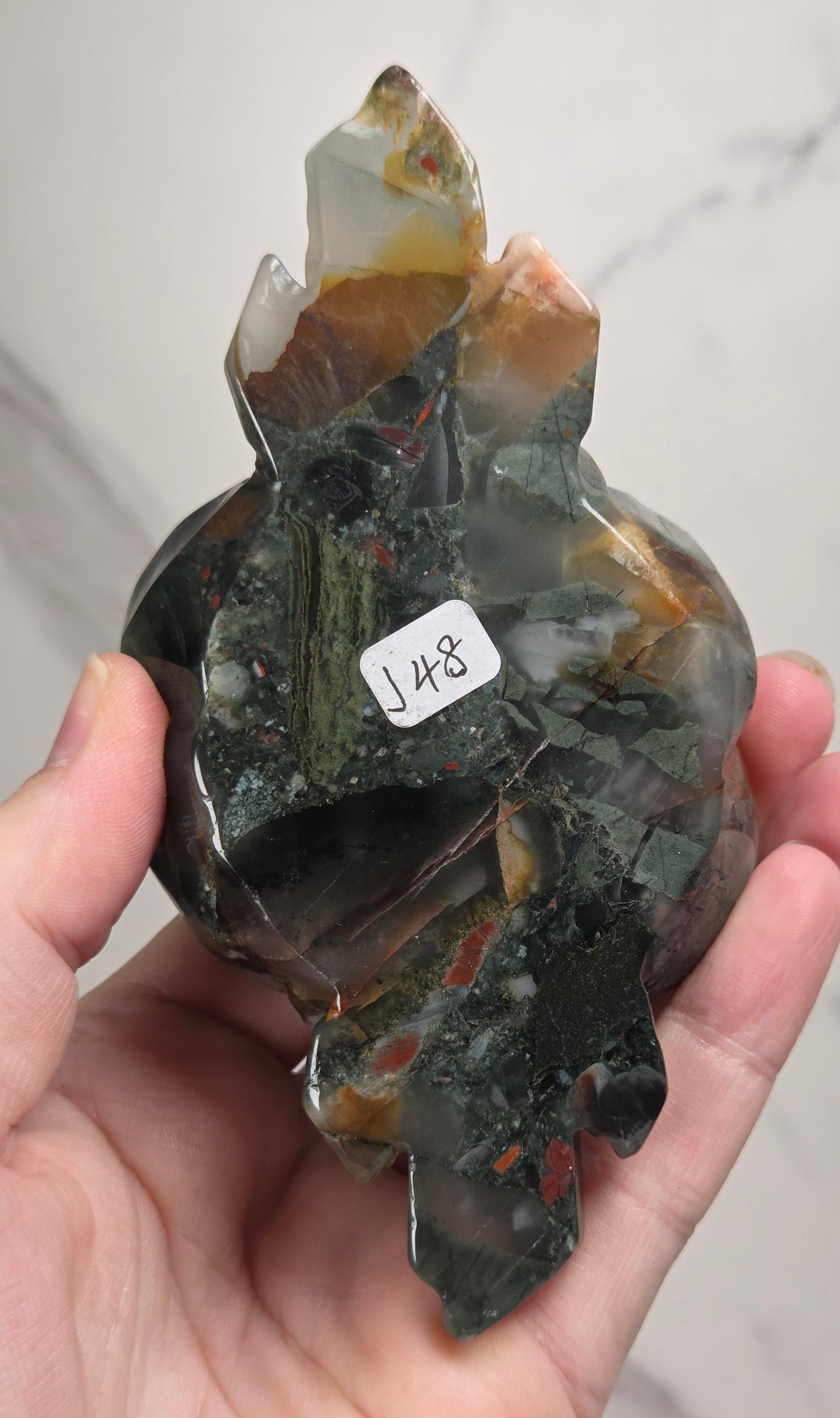 A hand holding a polished natural crystal cluster with multiple pointed mineral tips. The stone shows dark green, black, brown, and translucent gray areas with some orange-brown matrix.