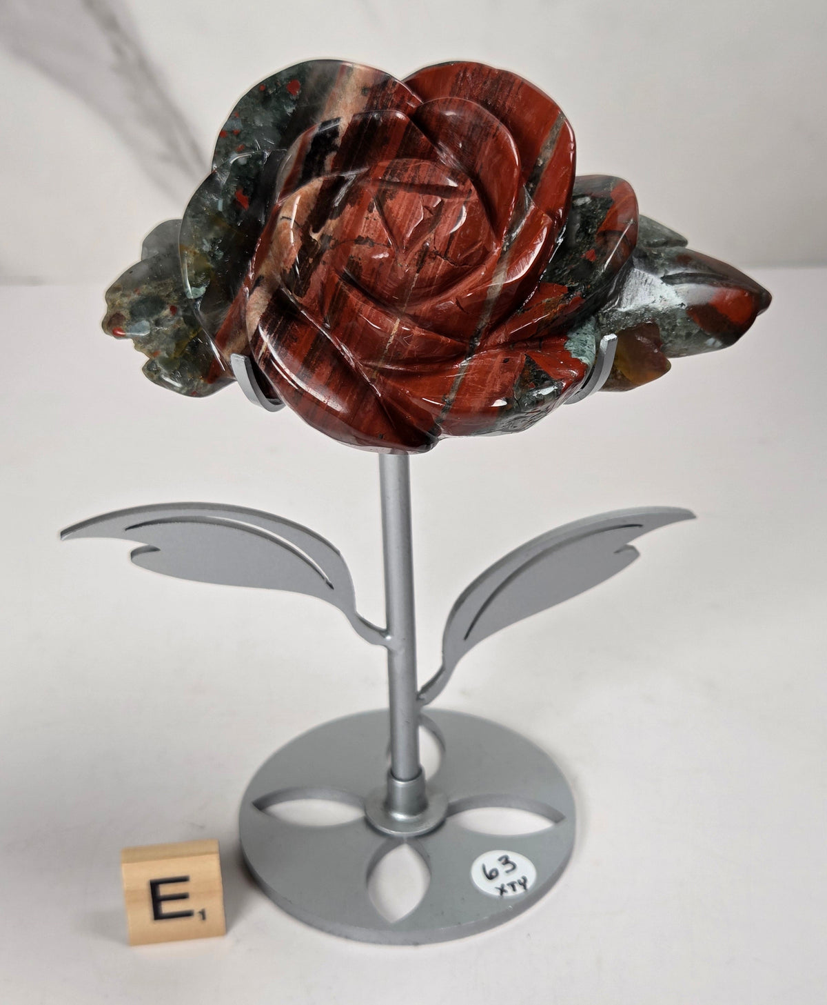 A carved rose sculpture made of reddish-brown and dark green stone, mounted on a silver metal stem with leaves and a round base, photographed on a white surface against a light gray marble-pattern background.