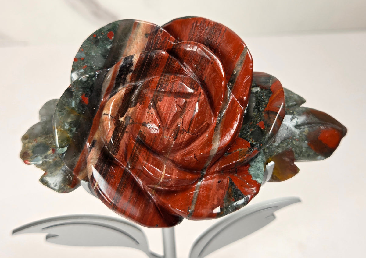 A carved rose-shaped decorative object made from polished red jasper stone with natural banding, displayed on a small metallic stand against a light neutral background.