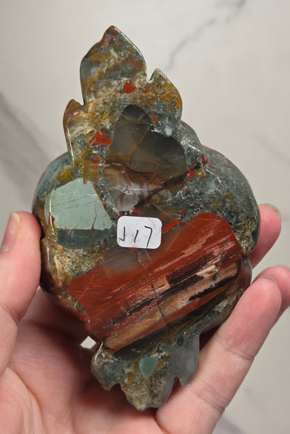 A carved stone rose sculpture with red, brown, and gray marbled petals on a silver metal stem and round base, displayed on a white surface with a small wooden letter tile nearby.