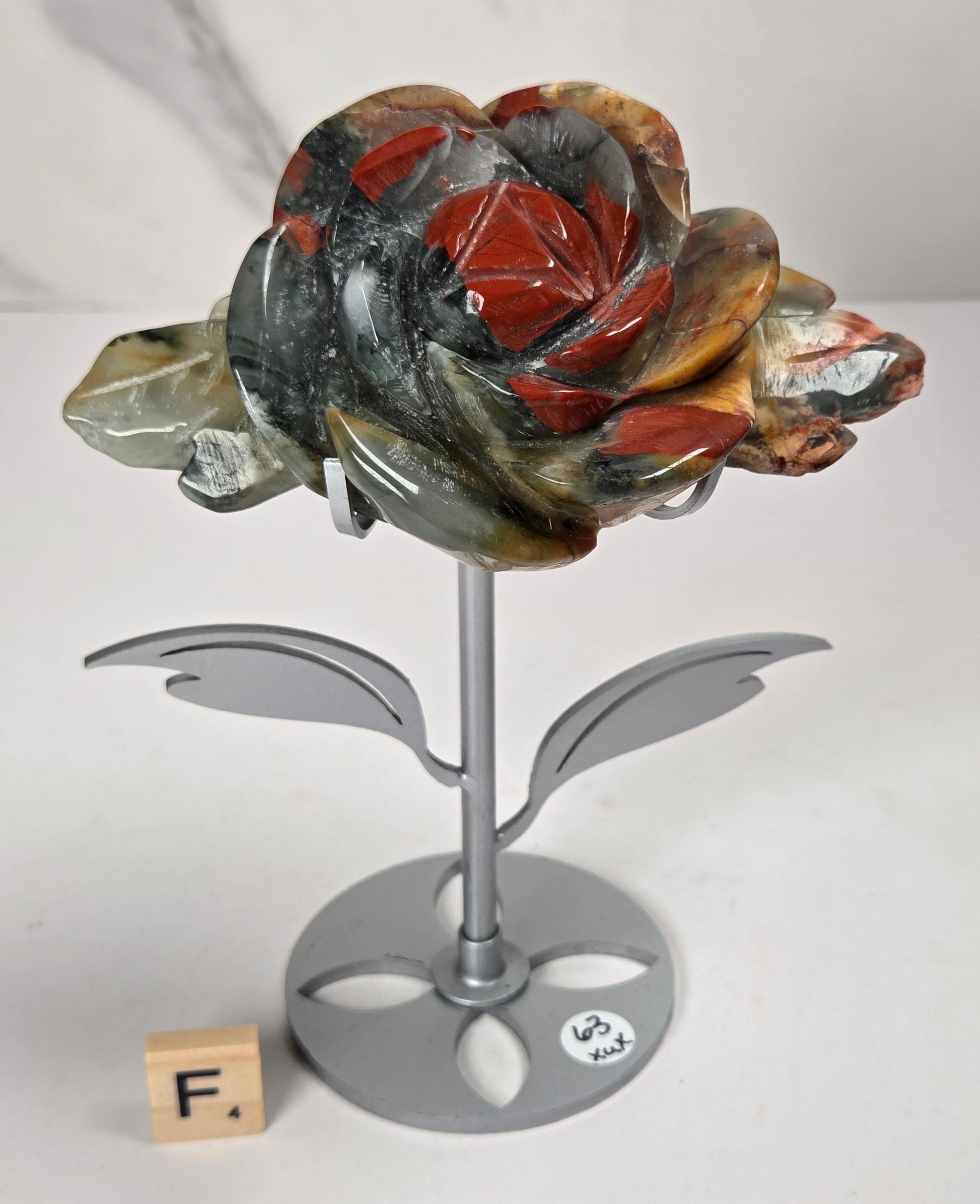 A carved stone rose sculpture with red, brown, and gray marbled petals on a silver metal stem and round base, displayed on a white surface with a small wooden letter tile nearby.
