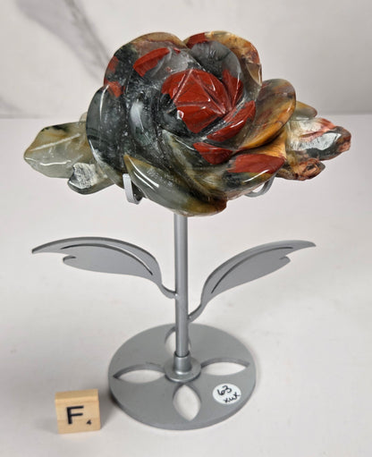 A carved stone rose sculpture with red, brown, and gray marbled petals on a silver metal stem and round base, displayed on a white surface with a small wooden letter tile nearby.