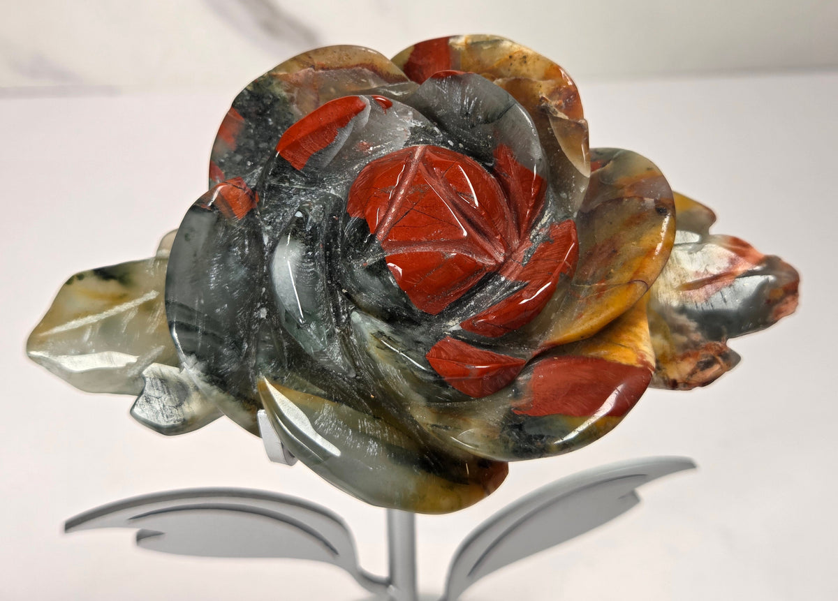 A carved rose-shaped decorative object made from polished stone with layered red, gray, black, and translucent amber tones, displayed on a small metallic stand against a light neutral background.