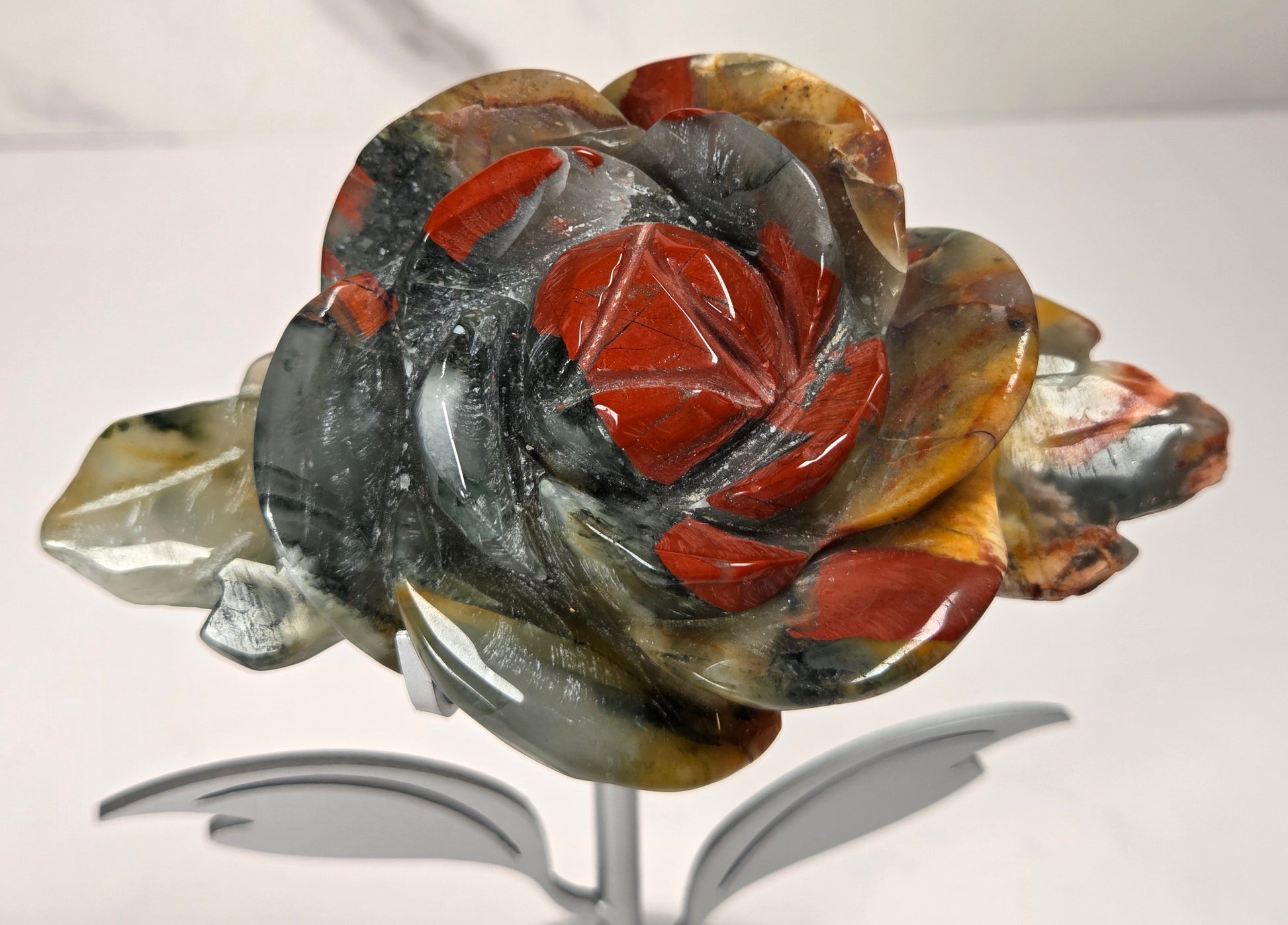 A carved rose-shaped decorative object made from polished stone with layered red, gray, black, and translucent amber tones, displayed on a small metallic stand against a light neutral background.