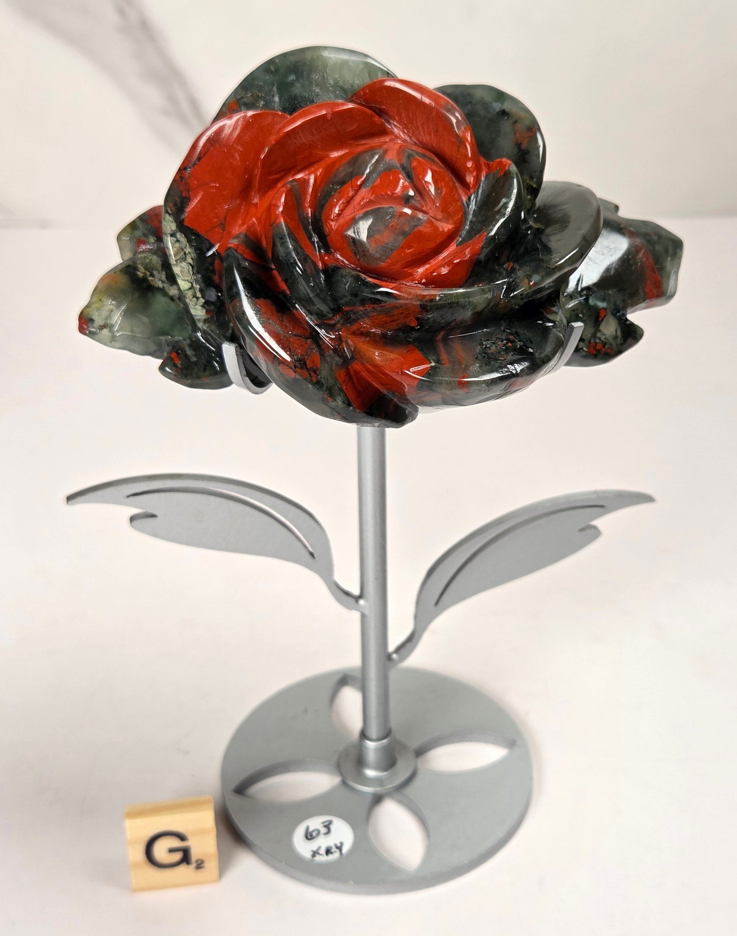A decorative sculpted rose with glossy red and dark gray marbled petals on a silver metal stand, photographed on a white surface against a light marble-patterned background.