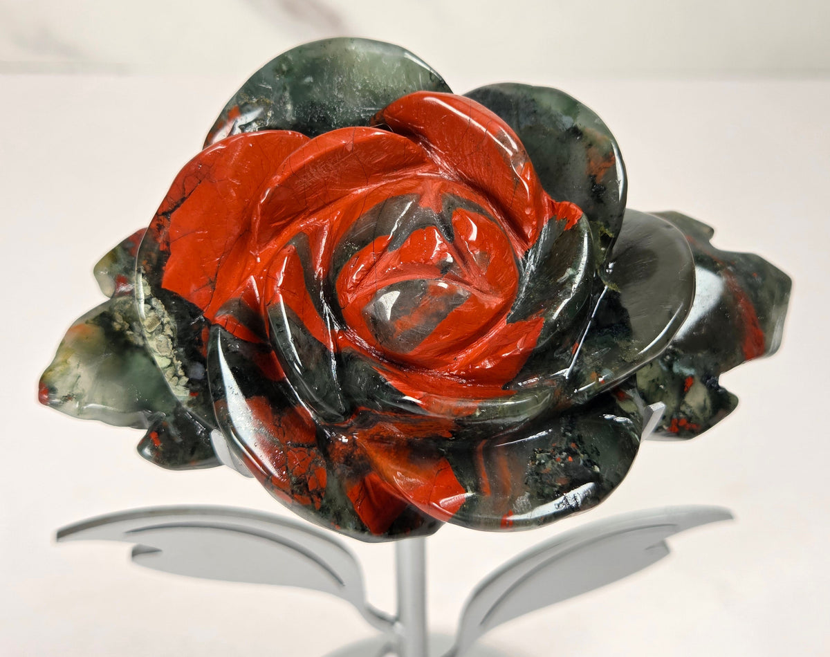 A carved stone rose sculpture with red, brown, and gray marbled petals on a silver metal stem and round base, displayed on a white surface with a small wooden letter tile nearby.