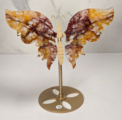 A decorative butterfly wall sculpture with marbled amber, brown, and cream wings mounted on a gold-toned metal stand against a white marble-patterned background.