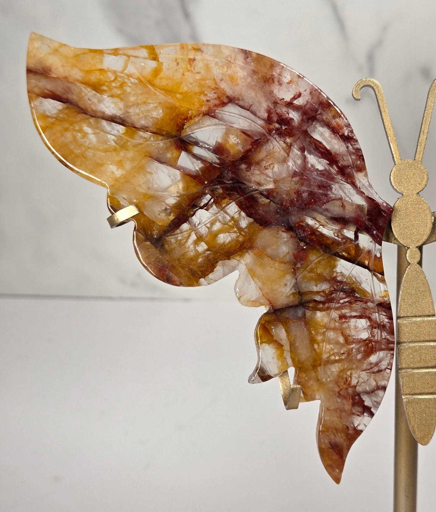 A decorative butterfly wall sculpture with marbled amber, brown, and cream wings mounted on a gold-toned metal stand against a white marble-patterned background.