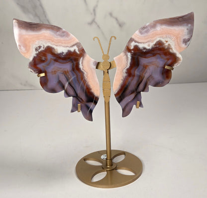 A butterfly-shaped jewelry display stand made from agate-patterned wings in shades of purple, brown, and peach, mounted on a gold-tone metal stand with a round base.