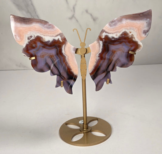 A butterfly-shaped jewelry display stand made from agate-patterned wings in shades of purple, brown, and peach, mounted on a gold-tone metal stand with a round base.