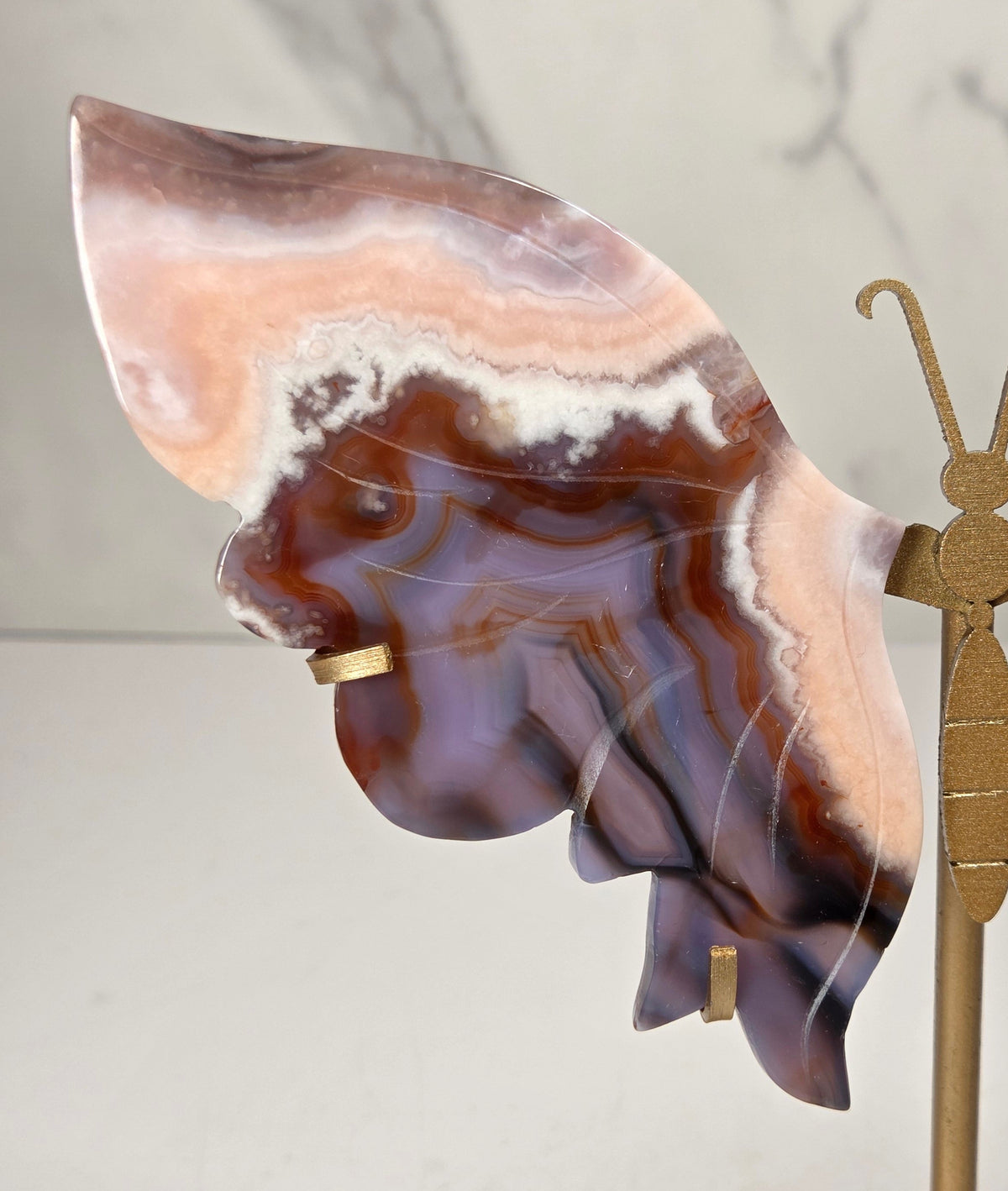 A decorative butterfly-shaped wall art piece made from polished agate stone with swirling brown, cream, and purple patterns, mounted on a gold-toned metal stand against a light marble-patterned background.