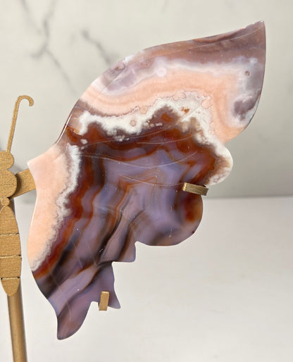 A butterfly-shaped jewelry display stand made from agate-patterned wings in shades of purple, brown, and peach, mounted on a gold-tone metal stand with a round base.