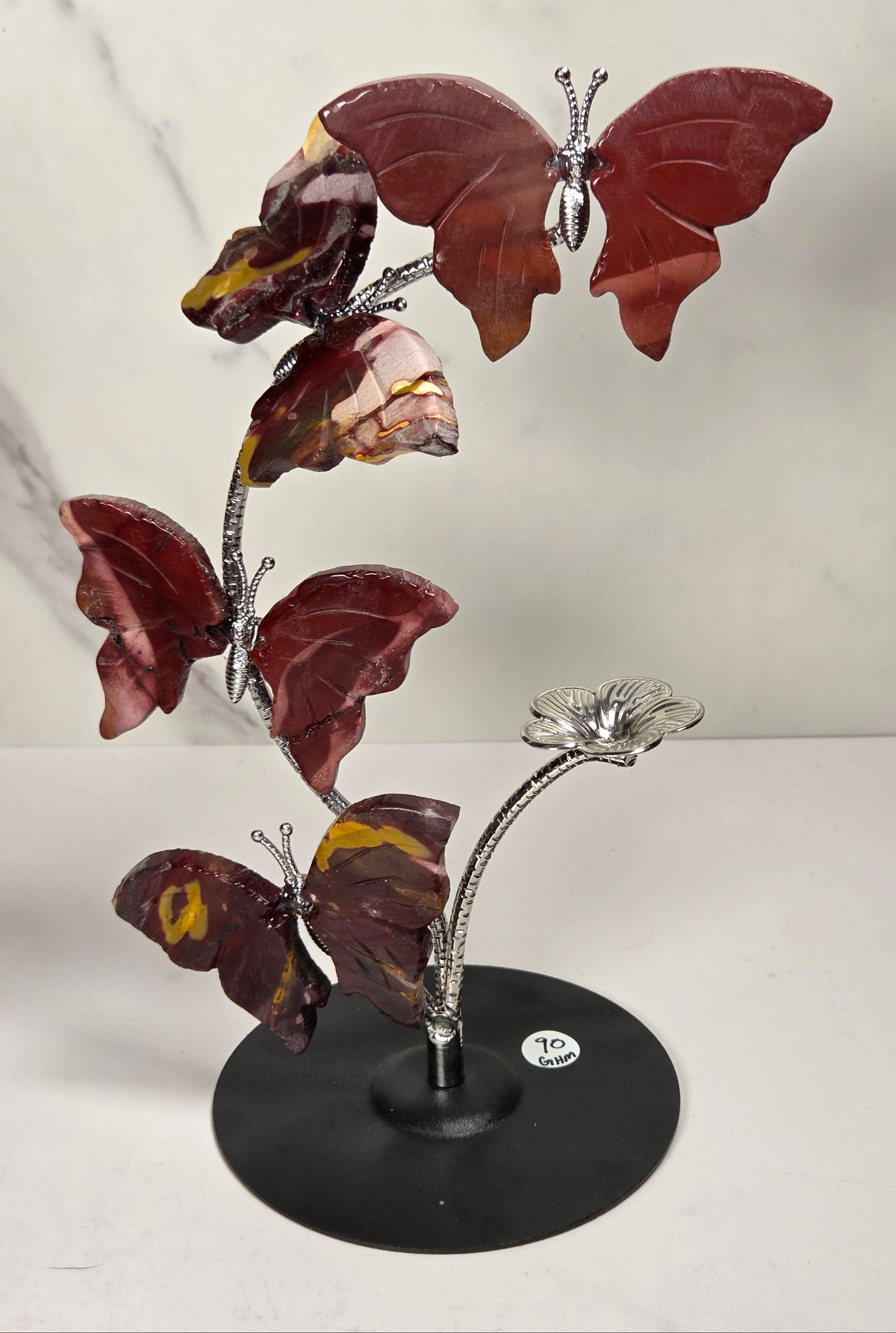 A decorative metal sculpture of four butterfly-like figures arranged along a flowering stem on a round black base, photographed against a light marble-pattern backdrop.