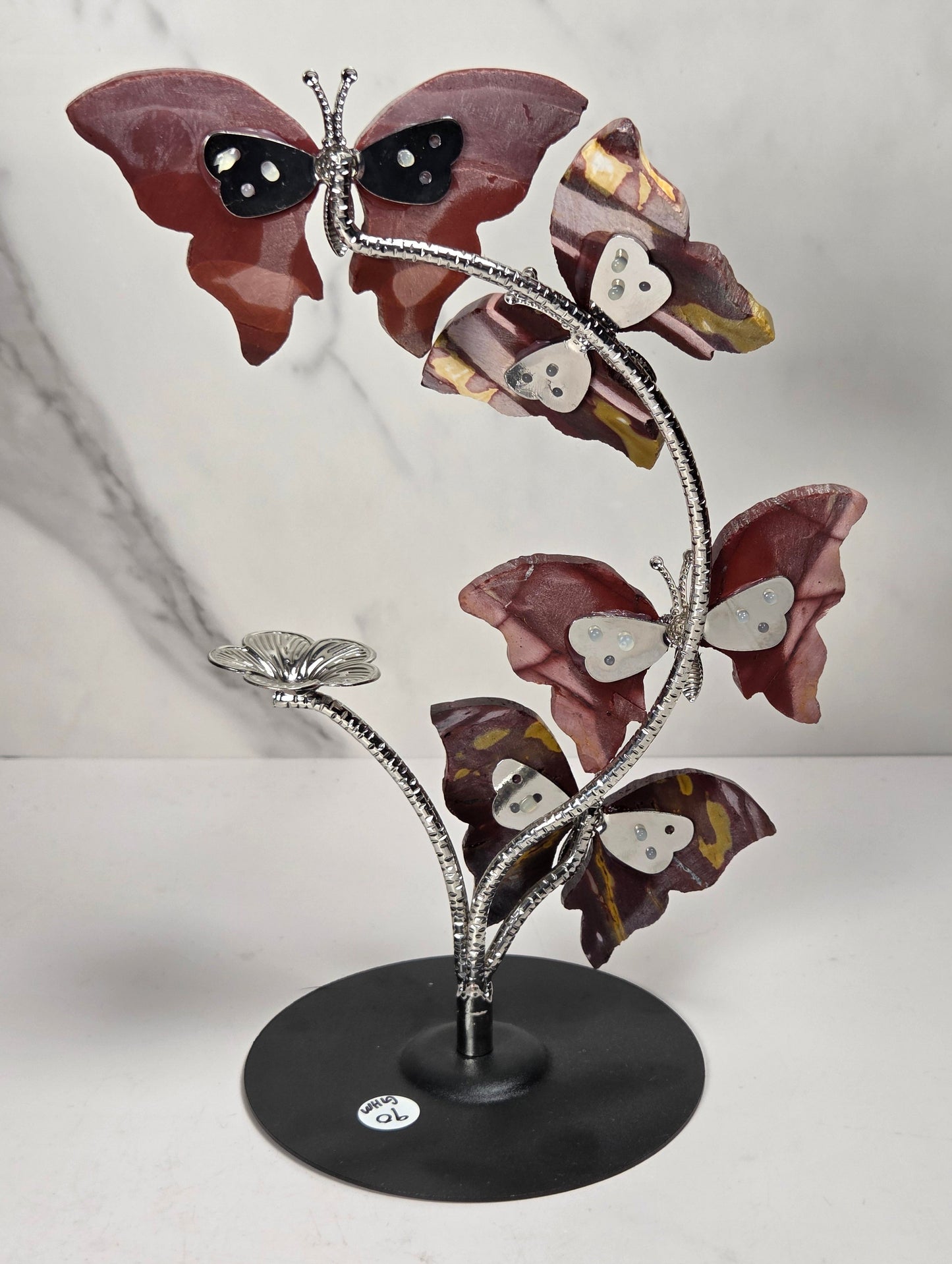 A decorative metal sculpture of four butterfly-like figures arranged along a flowering stem on a round black base, photographed against a light marble-pattern backdrop.