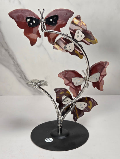 A decorative metal sculpture of four butterfly-like figures arranged along a flowering stem on a round black base, photographed against a light marble-pattern backdrop.