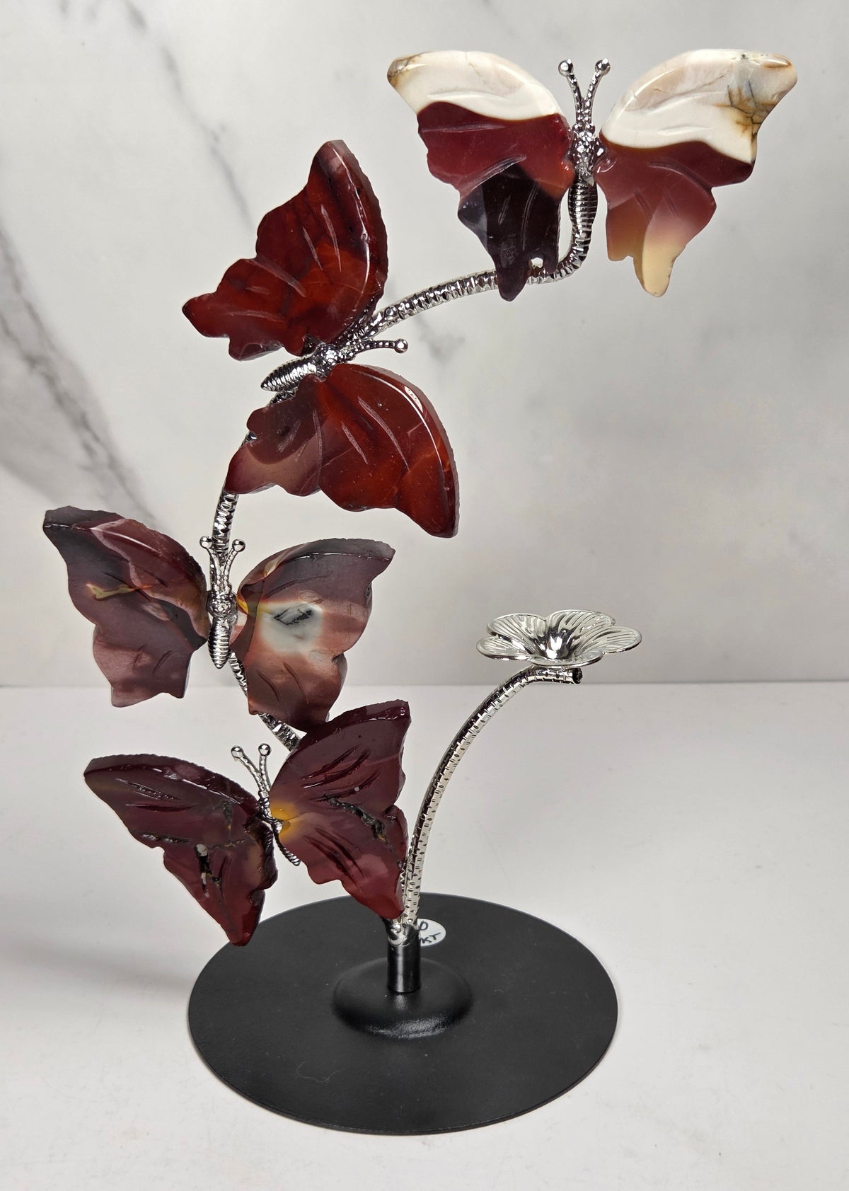 A decorative metal sculpture of four butterfly-like figures arranged along a flowering stem on a round black base, photographed against a light marble-pattern backdrop.