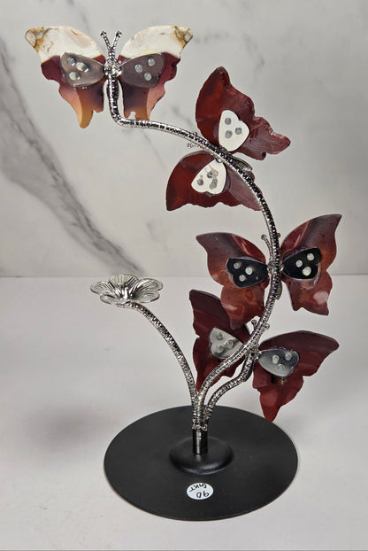 A decorative metal sculpture of four butterfly-like figures arranged along a flowering stem on a round black base, photographed against a light marble-pattern backdrop.