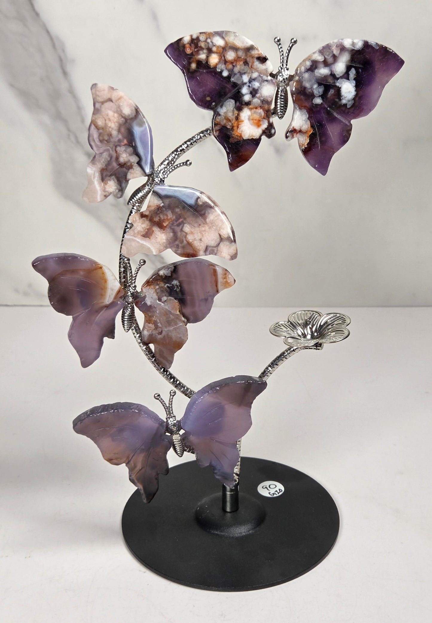 A decorative metal sculpture of four butterfly-like figures arranged along a flowering stem on a round black base, photographed against a light marble-pattern backdrop.