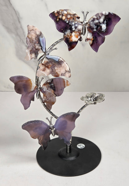 A decorative metal sculpture of four butterfly-like figures arranged along a flowering stem on a round black base, photographed against a light marble-pattern backdrop.