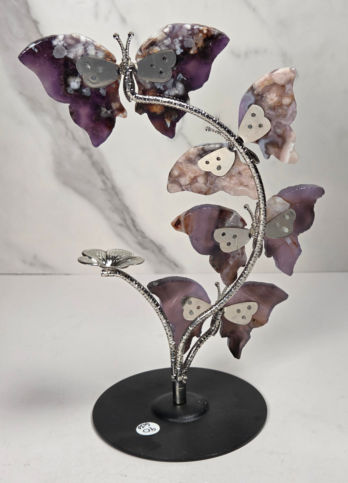 A decorative metal sculpture of four butterfly-like figures arranged along a flowering stem on a round black base, photographed against a light marble-pattern backdrop.