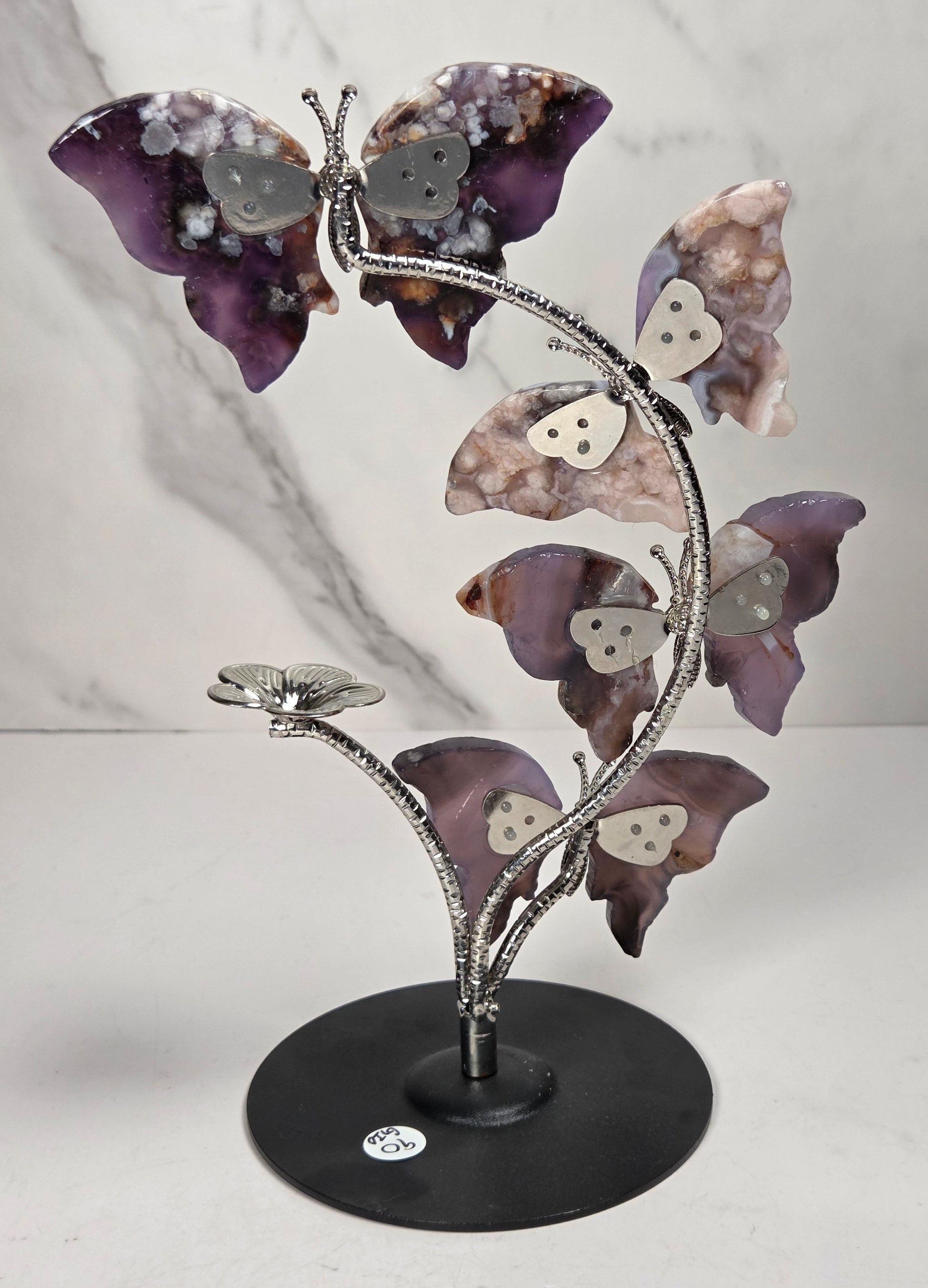 A decorative metal sculpture of four butterfly-like figures arranged along a flowering stem on a round black base, photographed against a light marble-pattern backdrop.