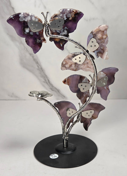 A decorative metal sculpture of four butterfly-like figures arranged along a flowering stem on a round black base, photographed against a light marble-pattern backdrop.