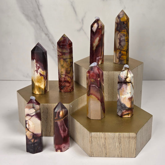 Seven polished crystal point towers with marbled patterns of brown, purple, gold, and cream, displayed on a tiered gold and wood geometric stand against a light marble background.