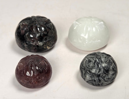 Four small carved stone beads or pendants arranged in a square on a plain light background. The beads are translucent and come in four colors: black, white, dark reddish-brown, and dark gray with marbled swirls.
