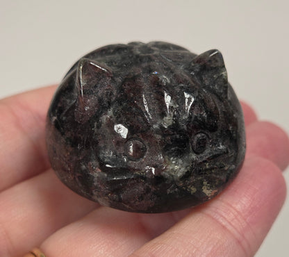 A hand holding a small polished dark cat-shaped stone or crystal with pointed ears and carved facial features, against a plain light background.
