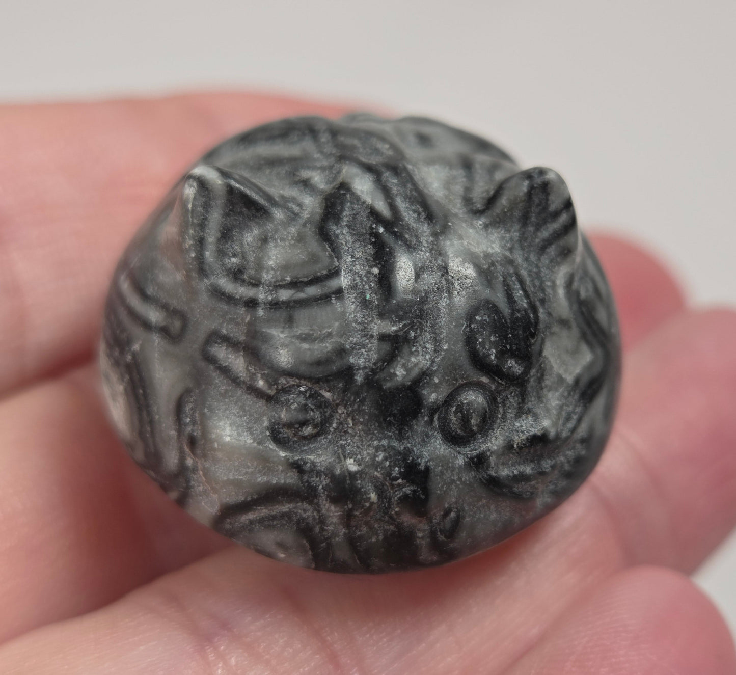 A small carved stone bead or ornament with an animal face motif, held in a person's hand against a soft light gray background.