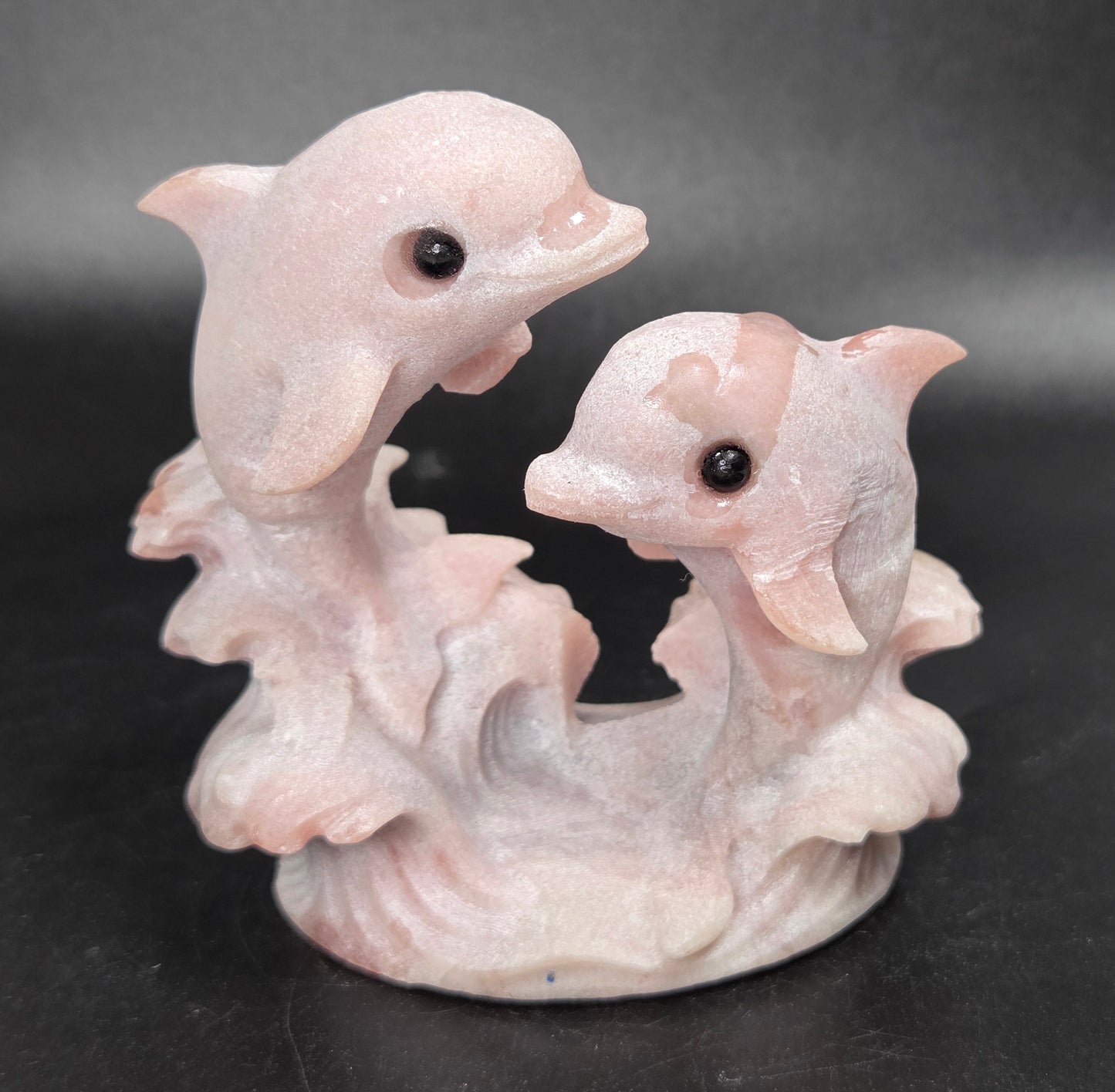 A pale pink sculpted figurine of two dolphins rising from stylized waves on a dark black background.