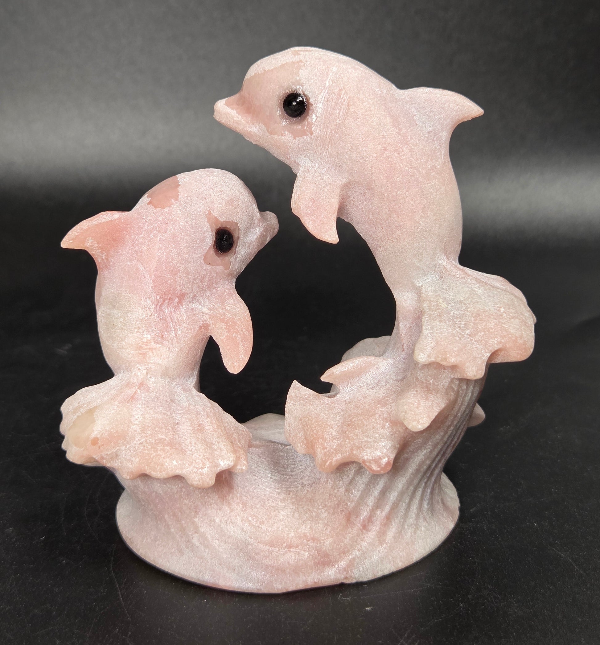 A pale pink carved figurine of two dolphins rising from stylized waves on a dark black background.
