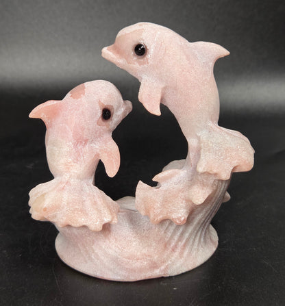 A pale pink carved figurine of two dolphins rising from stylized waves on a dark black background.