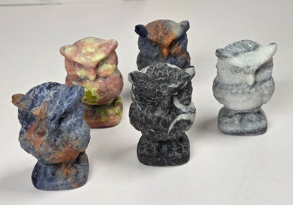 Five small carved animal figurines, including a blue-gray beaver, a dark gray boar, a pinkish owl, and others in earthy tones, arranged on a white surface against a light gray marble-patterned background.