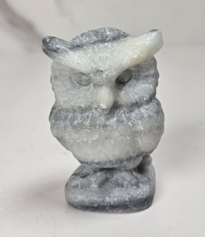 A small carved owl figurine made from natural stone, featuring a textured, speckled pattern of gray and off-white. The owl is depicted perched on a matching stone base against a neutral, light-colored background.