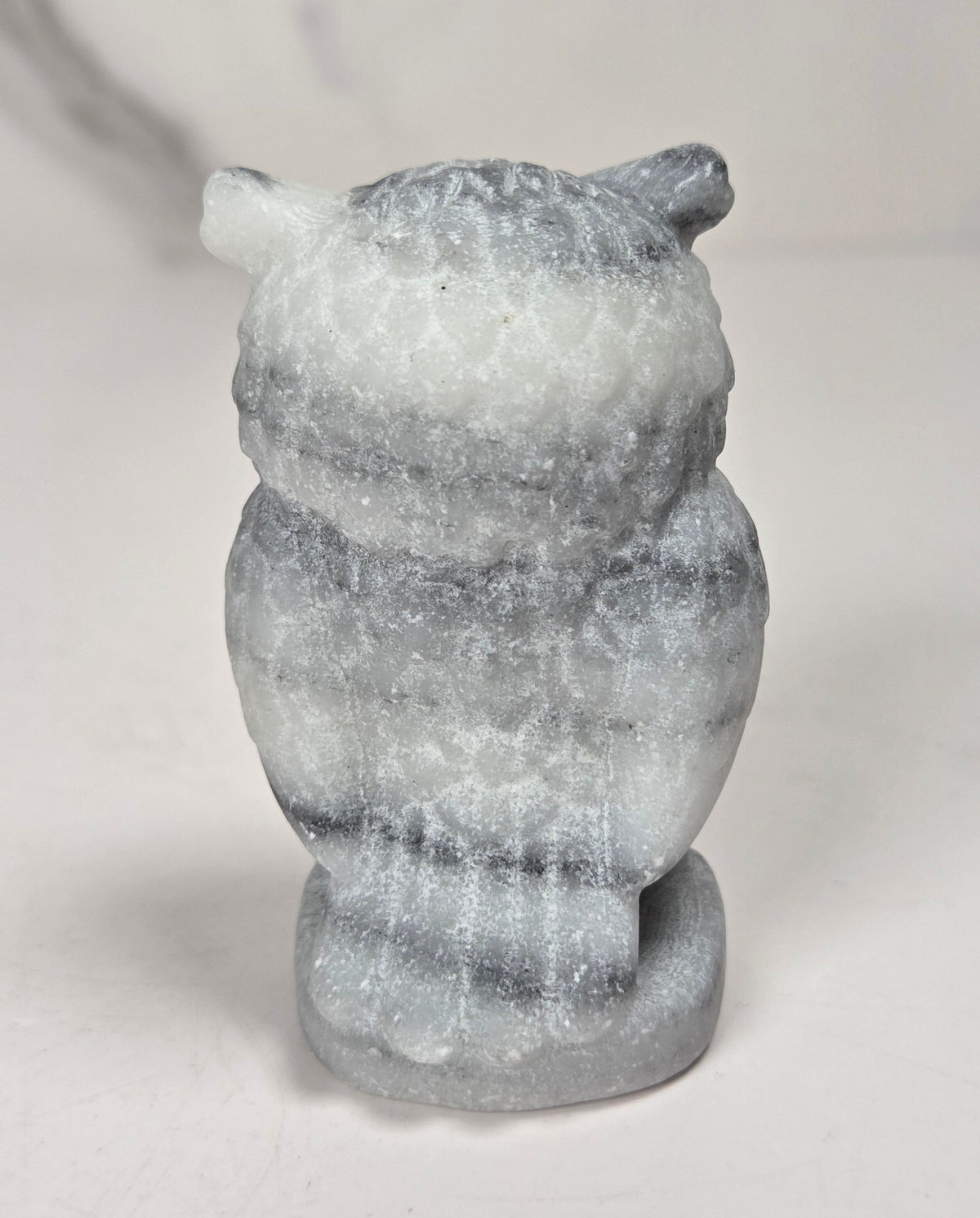 A small carved owl figurine made from layered gray and white stone, shown from the back on a light gray surface.
