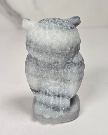 A small carved owl figurine made from layered gray and white stone, shown from the back on a light gray surface.