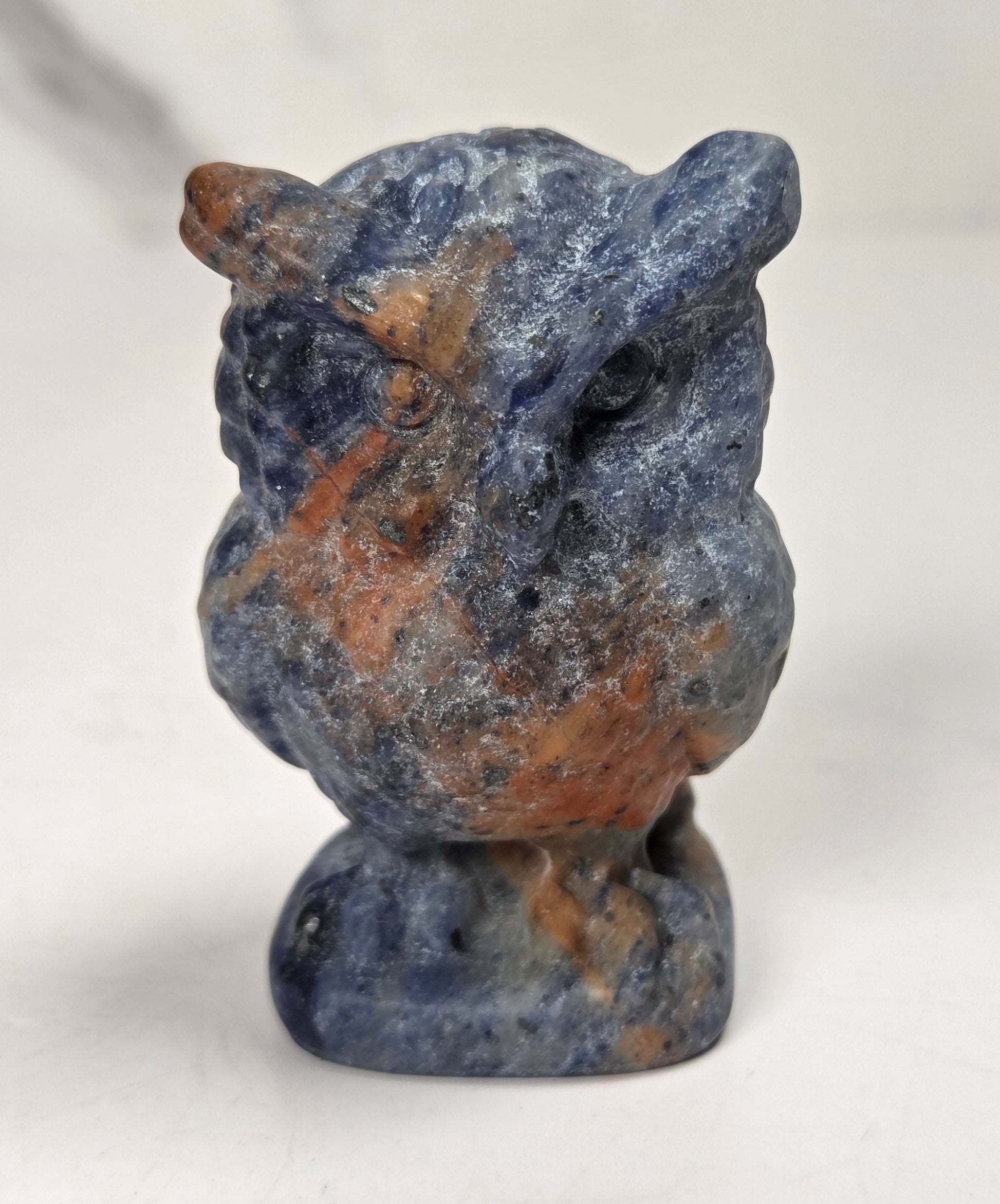 A small carved owl figurine made of blue lapis lazuli stone with prominent orange-brown copper inclusions, standing on a matching stone base against a light gray background.