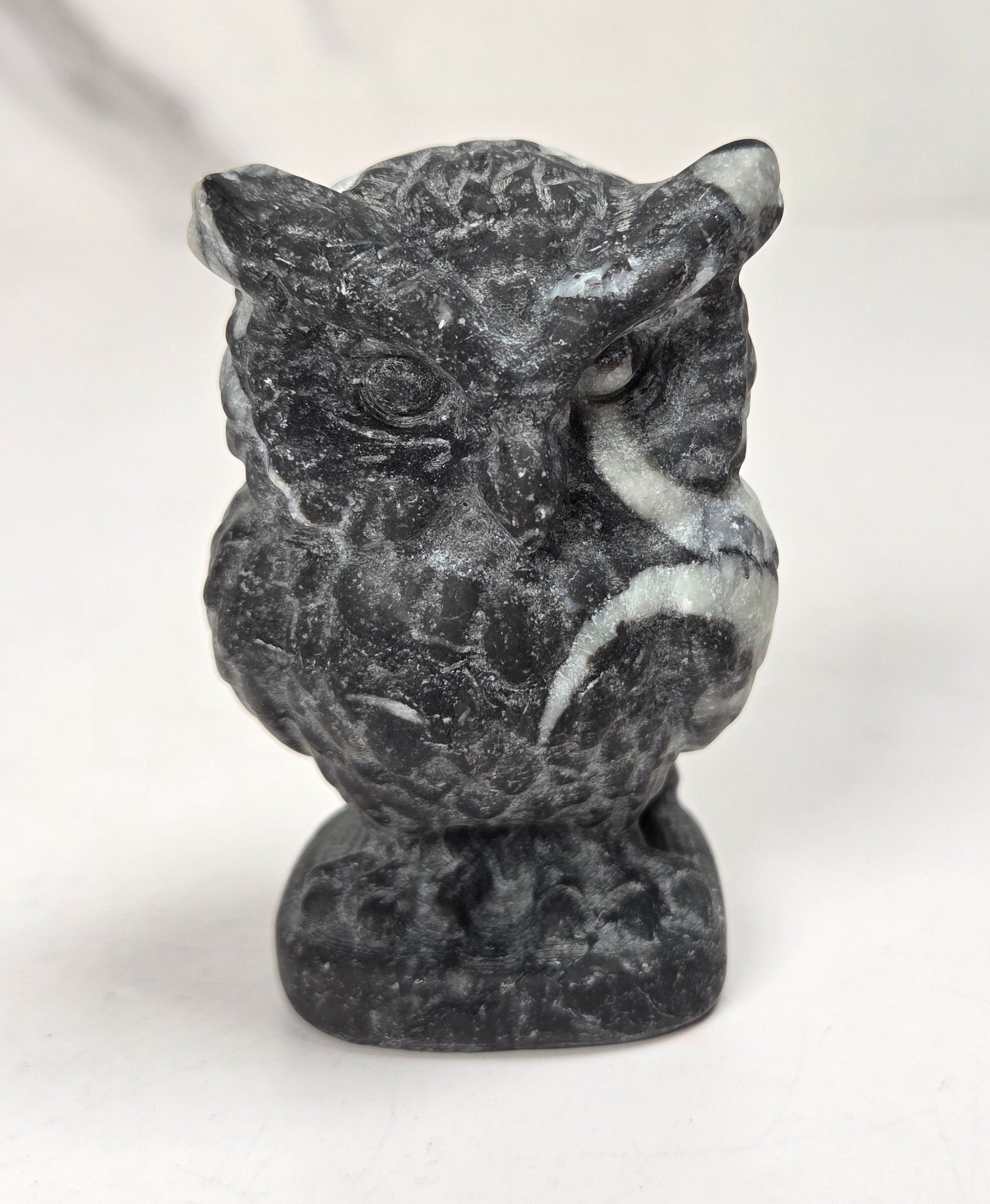 A small carved owl figurine made of dark gray stone with natural white veining, standing on a matching stone base against a light gray background.