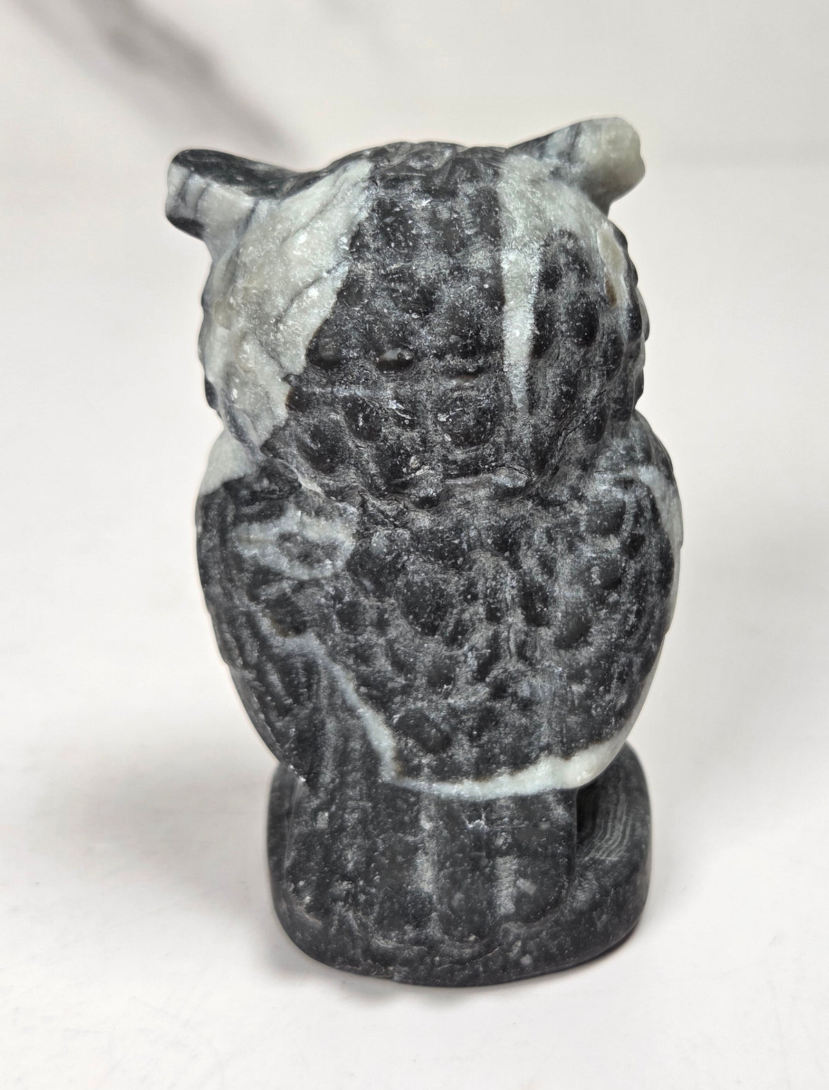 A small carved stone owl figurine, likely made of dark gray basalt or charcoal with white marbled streaks, shown from the back on a light gray surface.