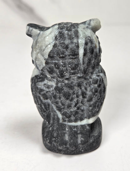 A small carved stone owl figurine, likely made of dark gray basalt or charcoal with white marbled streaks, shown from the back on a light gray surface.