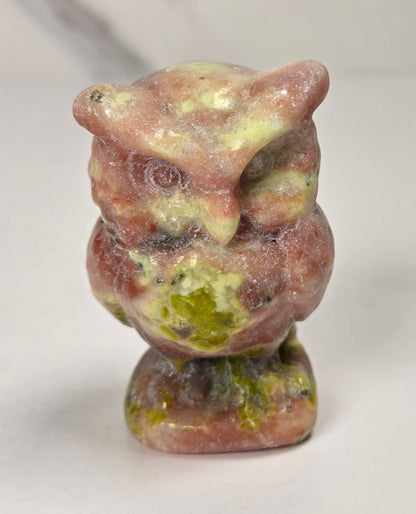 A small carved owl figurine made of pinkish stone with green and yellowish patina, standing on a white surface against a light gray background.